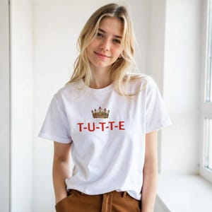 Könnte beinhalten: Weißes T-Shirt mit kurzen Ärmeln, roter und goldener Kronengrafik und dem Wort "T-U-T-T-E" in Rot. Das T-Shirt besteht aus einem weichen Material.