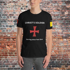 Puede incluir: Camiseta negra con la frase "CHRIST'S SOLDIER" sobre una cruz roja. Debajo, se lee "Serving since Year Zero". La camiseta tiene cuello redondo y mangas cortas.