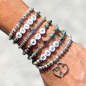 May include: A collection of beaded bracelets in silver and green. Some bracelets spell out "KYLER" and "MAMA" with white letter beads. Other bracelets feature heart and cross charms. One bracelet has a double heart charm.