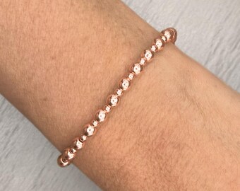 May include: Rose gold beaded bracelet. The bracelet features small, evenly spaced, round beads. The beads are strung together to form a flexible band. The bracelet is shown on a person's arm.