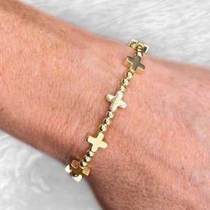 May include: Gold-colored bracelet featuring cross charms and small, round beads. The bracelet is worn on a wrist, showcasing the jewelry's design and color. The crosses are evenly spaced along the bracelet.