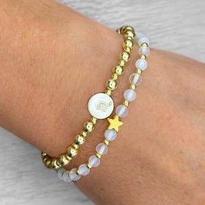 Op de afbeelding: Twee gouden kralenarmbanden. Eén armband heeft een witte cirkelvormige bedel met een gouden letter "m". De andere armband heeft kleine, heldere kralen en een gouden sterbedel. De armbanden worden om een pols gedragen.