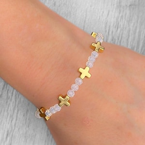 Peut inclure: Un bracelet doré orné de breloques en forme de croix et de petites perles transparentes. Le bracelet est porté au poignet, mettant en valeur le design délicat. Les croix sont espacées uniformément le long du bracelet.