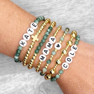 Pode incluir: Uma coleção de pulseiras de contas em vários estilos. Algumas pulseiras apresentam contas de letras brancas que soletram nomes como "KATE" e "COLE", enquanto outras têm contas douradas e contas de pedra verde. Uma pulseira inclui um berloque de cruz dourada.