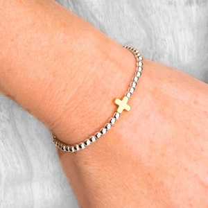 May include: A stretch bracelet composed of small, round, metallic silver-colored beads. The bracelet features a single, centered, gold-colored cross charm with a smooth, polished finish. The beads are uniform in size and arranged in a continuous circular loop.