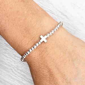 Op de afbeelding: Zilveren kralenarmband met een kleine, witte kruisbedel. De armband is gemaakt van kleine, ronde zilveren kralen. Het kruis is gecentreerd op de armband. De armband wordt om een pols gedragen.