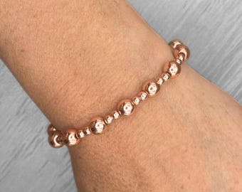 May include: Rose gold beaded bracelet. The bracelet features a series of round beads in varying sizes, strung together to form a flexible band. The beads have a shiny, metallic finish, reflecting light. The bracelet is worn on a wrist.
