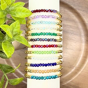 May include: A collection of beaded bracelets in various colors, including red, purple, blue, and green. Each bracelet features small, colorful beads and gold-colored accents. The bracelets are displayed on a white cylindrical object.