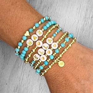May include: Stack of personalized bracelets. The bracelets feature turquoise beads, gold-colored beads, and white letter beads spelling out "MAX" and "ELLA". Some bracelets have heart charms.