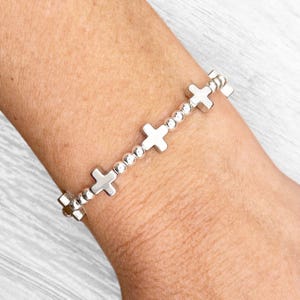 May include: A silver bracelet featuring a series of cross charms interspersed with small, round beads. The bracelet is worn on a wrist, showcasing its design and the reflective quality of the metal. The crosses are evenly spaced, creating a balanced and elegant look.