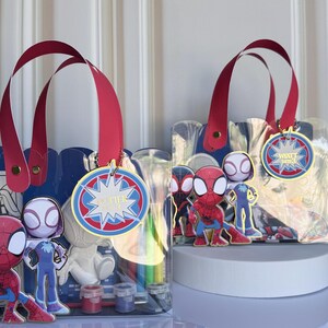 May include: Two clear tote bags with red handles and Spider-Man themed decorations. Each bag features a blue scalloped top, a round tag with text, and various Spider-Man character figures. The bags contain art supplies.