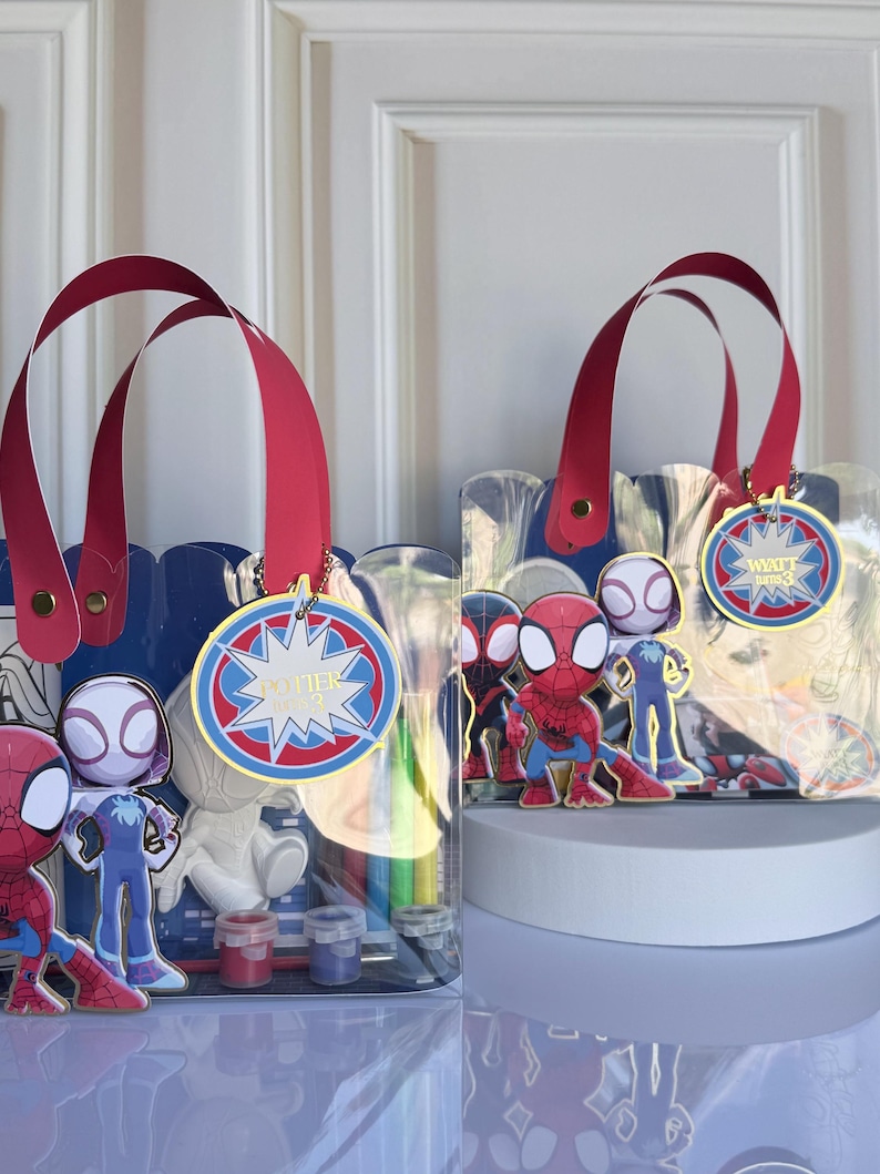 May include: Two clear tote bags with red handles and Spider-Man themed decorations. Each bag features a blue scalloped top, a round tag with text, and various Spider-Man character figures. The bags contain art supplies.