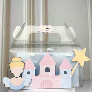 Puede incluir: Una caja de regalo transparente con temática de princesa. La caja contiene una ilustración de castillo rosa y azul, un recorte de Cenicienta y una varita mágica amarilla en forma de estrella. La caja tiene un asa y una etiqueta que dice "ana Couple".