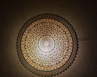 May include: Ornate, circular light fixture with a detailed, openwork design. The fixture has a central light source, casting intricate shadows on the surrounding dark surface. The design features floral and geometric patterns.