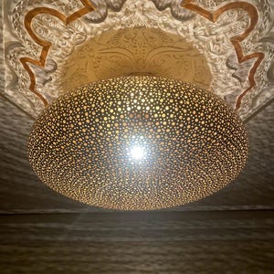 Può includere: Un lampadario decorativo a soffitto con un paralume sferico perforato. La luce emana dal centro del paralume. Il soffitto sopra la lampada presenta intagli ornati e dettagli dorati.