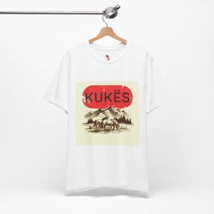 May include: White t-shirt with a red oval design displaying the word "KUKËS". The graphic below shows mountains, trees, and two horses. The t-shirt is hanging on a wooden hanger.