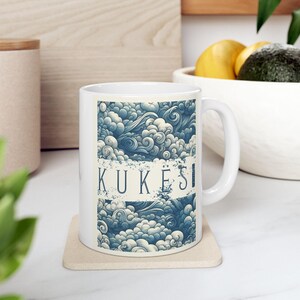 May include: White ceramic mug with a blue and white cloud pattern and the word "KUKESI". The mug has a curved handle and is on a beige coaster. The design evokes a sense of calm.