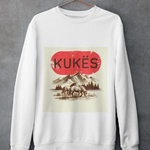 May include: White sweatshirt with a graphic featuring the word "KUKËS" in a red oval, with a mountain and horse illustration. The design is in shades of brown and tan.
