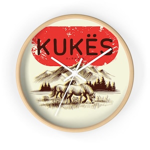 May include: A round wall clock with a light wood frame. The clock face features a red oval with the word "KUKËS" in dark letters, and a drawing of two horses grazing in a field with mountains in the background. The word "ALBANIA" is below "KUKËS".