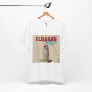 May include: White t-shirt with a graphic of the Elbasan Clock Tower in Albania. The design includes the text "ELBASAN" in red and "Albania" in blue. The shirt is displayed on a wooden hanger.
