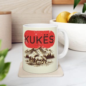 May include: White ceramic mug with the word "KUKËS" in black letters inside a red oval, with "ALBANIA" below. The mug design features a mountain range and two horses grazing. Ideal for tea or coffee. A great gift idea.