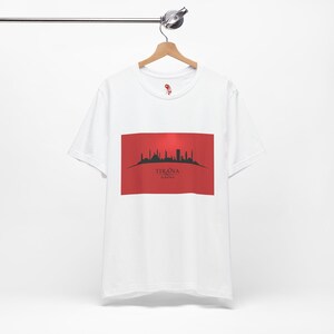Pode incluir: Camiseta branca com um design retangular vermelho com um horizonte preto e a palavra "TIRANA". A camiseta está pendurada em um cabide de madeira.