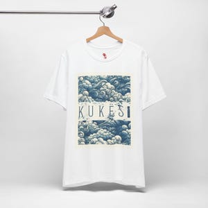 May include: White t-shirt with a rectangular graphic featuring a blue and white cloud design and the word "KUKËSI" in a serif font. The shirt is hanging on a wooden hanger.