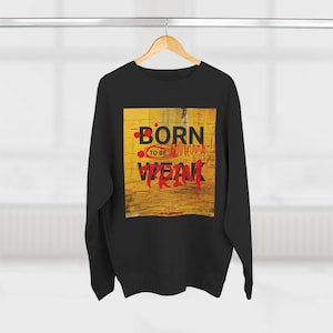 May include: Black sweatshirt with a graphic on the front. The graphic features the words "BORN TO BE WEAK" in red and black, set against a yellow and brown background. The sweatshirt is hanging on a clothing rack.