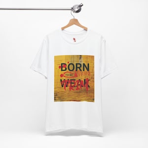 Może przedstawiać: Biała koszulka z żółto-brązową grafiką. Grafika głosi "BORN TO BE WEAK" czarnymi i czerwonymi literami. Koszulka wisi na drewnianym wieszaku.
