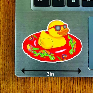 May include: A yellow rubber duck wearing red sunglasses floats in a red inner tube decorated with cartoon turtles and fish. The sticker measures 3 inches across.