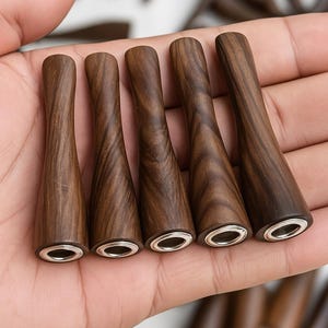 May include: Five wooden smoking pipe mouthpieces with a dark brown finish and silver-colored metal rings at the base. The wood grain is visible. Other similar pieces are in the background.