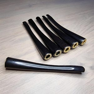 May include: A collection of six black pipe stems with brass-colored metal rings at the end. One stem is positioned in the foreground, showcasing the smooth, curved design. The stems are arranged on a light-colored wooden surface.