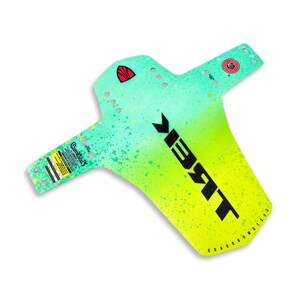 Peut inclure: Un garde-boue de vélo avec un dégradé vert et jaune, parsemé de peinture. Le mot "TREK" est imprimé en grandes lettres noires. Du texte et des logos supplémentaires sont visibles, dont "Cometag" et un emblème rouge.