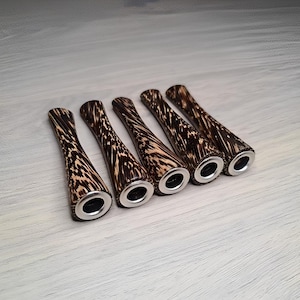 Può includere: Sei bocchini per pipa in legno con un motivo marrone e beige unico. Ogni bocchino ha una forma svasata e un'estremità in metallo argentato. Le pipe sono disposte in fila su una superficie di legno chiaro.