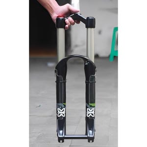May include: Black and silver bicycle suspension fork with a curved top and two parallel tubes. The fork has white and green graphics and is held by a hand at the top.