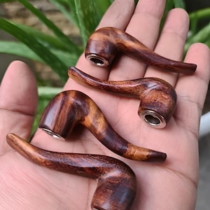 May include: Four handcrafted wooden pipes with a curved design are displayed in a hand. The pipes are made of dark brown wood with a polished finish and silver-colored metal accents. The pipes are a tobacco smoking accessory.
