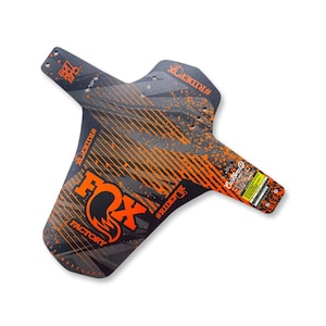 Peut inclure: Un garde-boue de vélo de montagne Fox Factory noir et orange. Il présente un motif géométrique, le logo Fox, et les mots "FACTORY" et "#RIDEFOX". Conçu pour protéger le vélo de la boue et des débris.