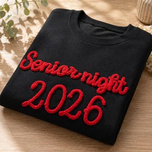 May include: Black sweatshirt with the words "Senior night 2026" in red, textured lettering. The sweatshirt is folded on a wooden surface. The design is a bold, eye-catching statement.