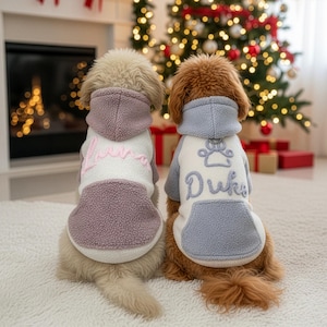 Puede incluir: Dos perros con suéteres de forro polar con capucha a juego. Uno es blanco y malva con el nombre "Luna" bordado en rosa. El otro es blanco y azul con "Duke" bordado en azul, con una huella de pata encima del nombre. Están sentados frente a un árbol de Navidad.