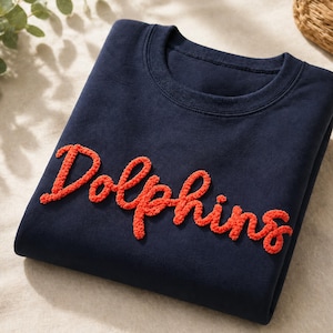 Könnte beinhalten: Ein marineblaues Sweatshirt mit dem Wort "Dolphins" in orangefarbener, strukturierter Schrift. Das Rundhals-Sweatshirt ist ordentlich gefaltet und zeigt den auffälligen Text. Ein schlichtes und sauberes Design für den Alltag.