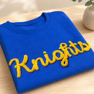 May include: A royal blue sweatshirt with the word "Knights" in yellow, textured lettering. The crew neck sweatshirt is made of a soft material and is displayed on a light wooden surface.