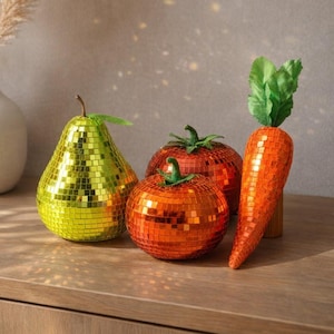 May include: A decorative still life featuring a pear, two tomatoes, and a carrot, all covered in mosaic tiles. A white vase with dried pampas grass is to the left. The pear is green, the tomatoes are red and orange, and the carrot is orange.