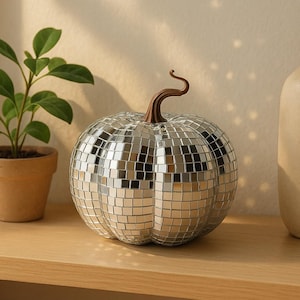 May include: A mirrored pumpkin ornament with a brown stem is displayed on a wooden shelf, alongside a small potted plant and a beige vase. This decorative pumpkin is covered in small, reflective squares, making it a striking home decor piece.