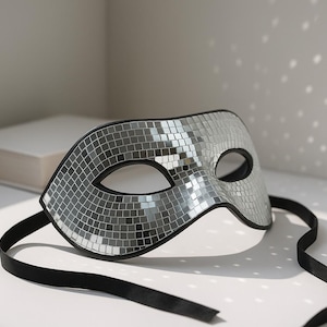 May include: A silver mosaic mask with black trim and eye openings. The mask has a black ribbon attached. The mask is resting on a white surface with a book in the background. The mask is likely for a masquerade or costume party.