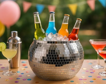 May include: A silver disco ball bowl filled with ice and four colourful bottles of drinks. Two cocktails with orange garnishes and a cocktail shaker are on a wooden table with confetti and balloons in the background.