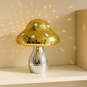 Pu&ograve; includere: Una lampada decorativa a forma di fungo con un cappello a mosaico dorato e una base a mosaico argentato. La lampada &egrave; su una mensola bianca con due libri che hanno le parole "UNION" e "HOME" sul dorso. La lampada proietta un motivo di luce sulla parete.