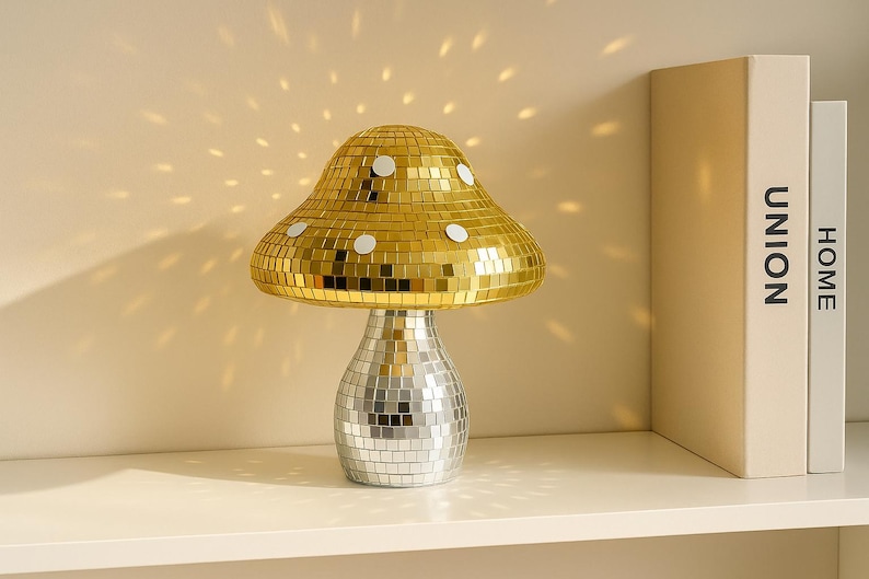 Pu&ograve; includere: Una lampada decorativa a forma di fungo con un cappello a mosaico dorato e una base a mosaico argentato. La lampada &egrave; su una mensola bianca con due libri che hanno le parole "UNION" e "HOME" sul dorso. La lampada proietta un motivo di luce sulla parete.