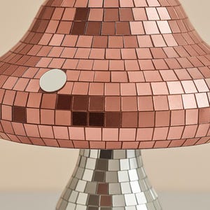 Pu&ograve; includere: Una lampada da tavolo a forma di fungo con un design a mosaico. La parte superiore della lampada &egrave; color rame e la base &egrave; argentata. La lampada ha un dettaglio circolare bianco sul lato.
