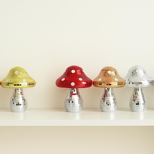 Pu&ograve; includere: Sei lampade decorative a forma di fungo con basi a sfera da discoteca e cappelli a mosaico colorati. I cappelli sono nei toni del rame, giallo, rosso, oro, argento e viola. Le lampade sono su una mensola bianca.