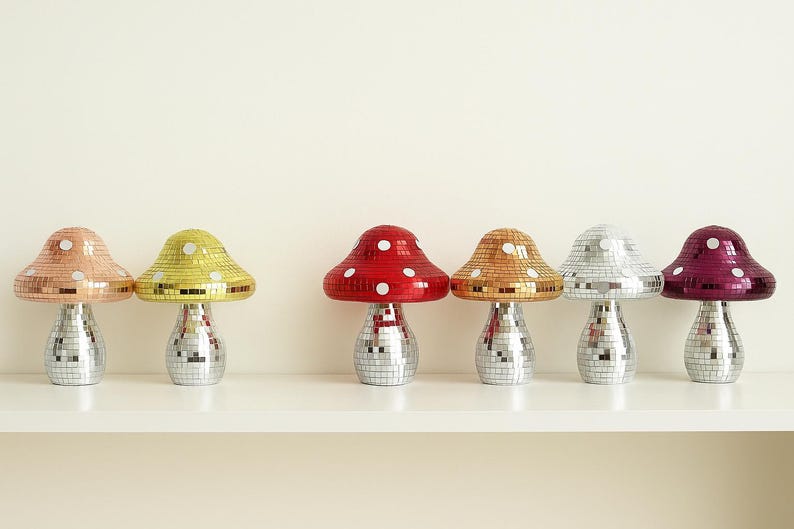Pu&ograve; includere: Sei lampade decorative a forma di fungo con basi a sfera da discoteca e cappelli a mosaico colorati. I cappelli sono nei toni del rame, giallo, rosso, oro, argento e viola. Le lampade sono su una mensola bianca.